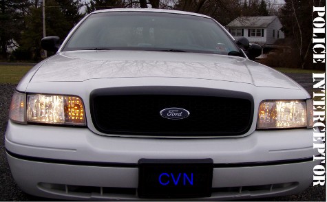 1999 Ford Crown Victoria - A white Ford Crown Victoria Police Interceptor is shown from the front, with a black grille and illuminated headlights.