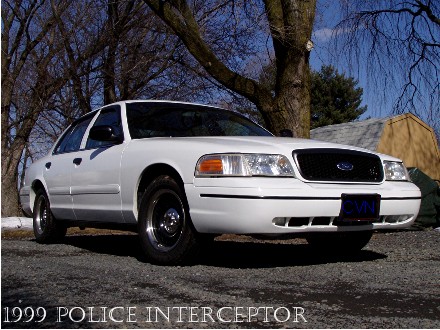 1999 Ford Crown Victoria - A white 1999 Ford Crown Victoria Police Interceptor is shown from a 3q-front angle, with text indicating the year and model visible at the…