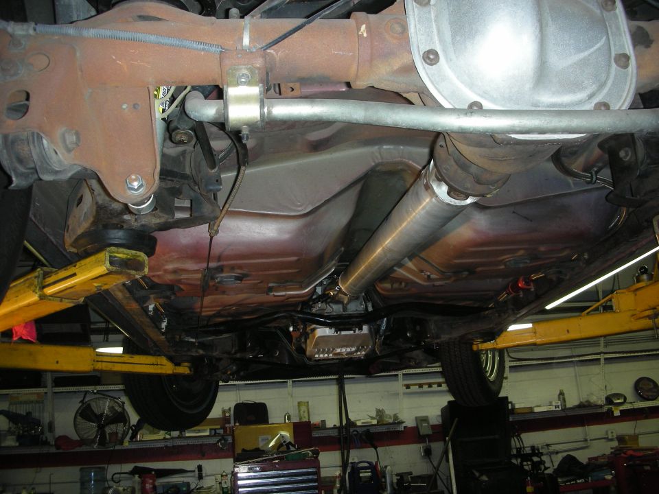 Undercarriage of a red vehicle showing the transmission, driveshaft, and exhaust system, lifted on a yellow car lift