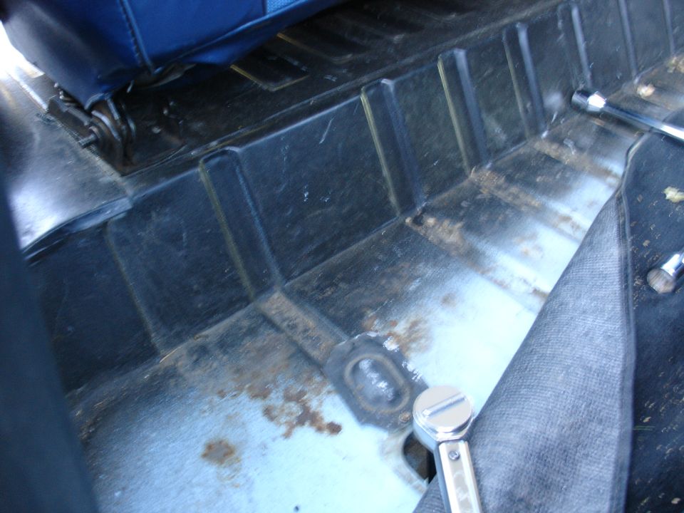 Bare ribbed floor pan with surface rust, grommet plug and chrome ratchet during interior teardown