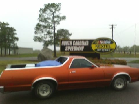 1979 Ford Ranchero - Side view of an orange 1979 Ford Ranchero pickup truck with a blue tarp in the bed, parked near a NASCAR sign.