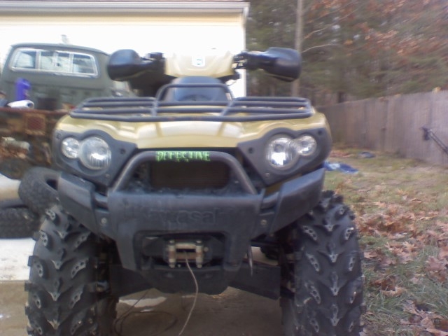 Front view of green and tan Kawasaki ATV with mud tires parked on grass by a fence.