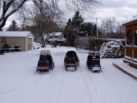 1999 Ford Crown Victoria - Three snowmobiles are parked in a snow-covered yard with houses and trees in the background.