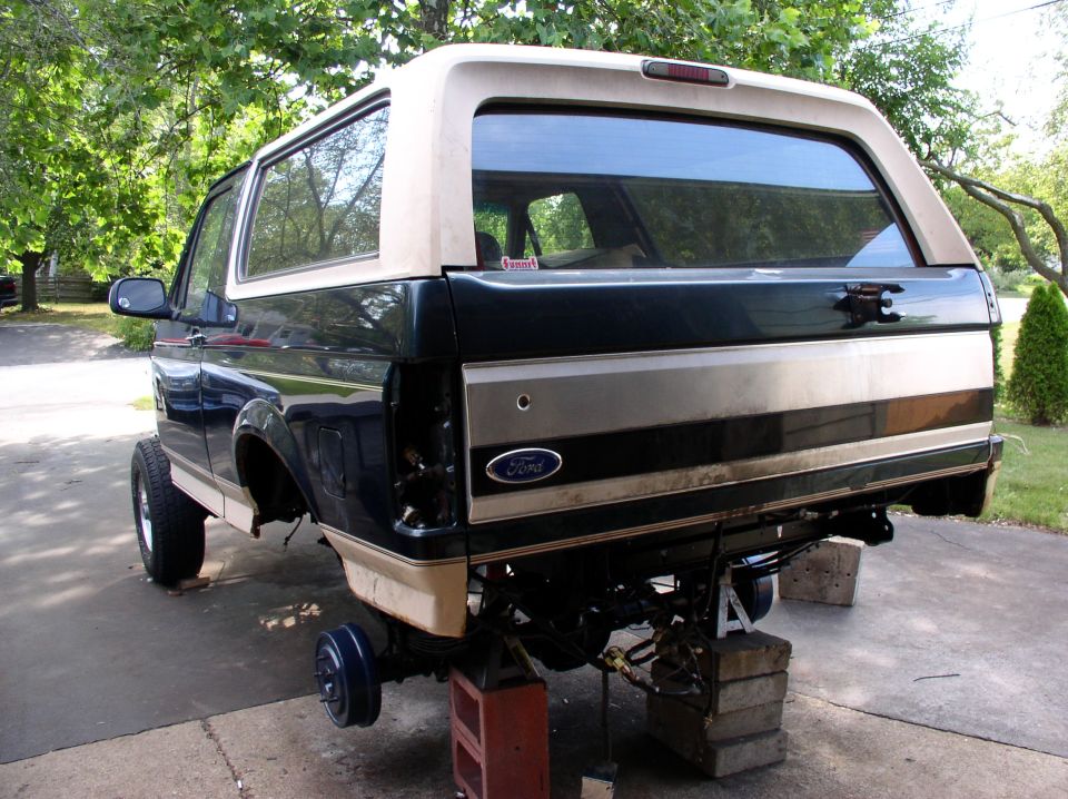 Green and tan Ford Bronco on cinder blocks in driveway, rear wheels and bumper removed