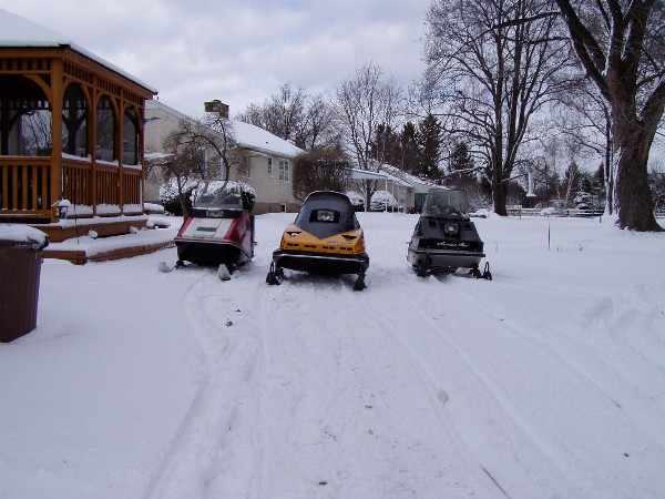 Three snowmobiles parked in a snowy yard next to a gazebo