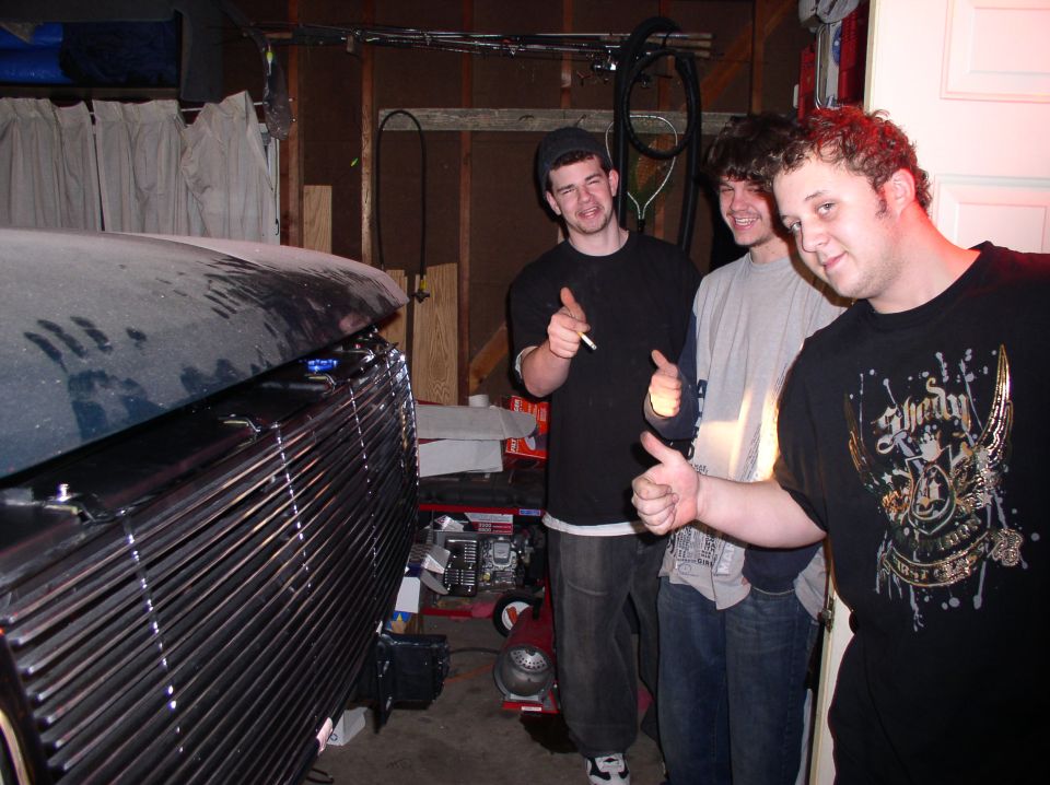 Car grille and three guys in a garage. One guy is smoking
