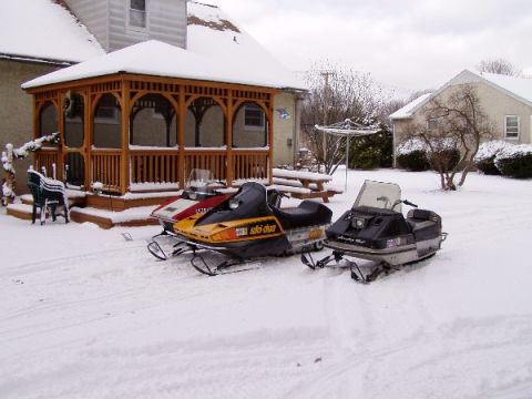 1999 Ford Crown Victoria - Two snowmobiles sit in the snow next to a wooden gazebo and a house.
