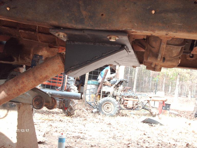 Custom bracket bolted to underside of vehicle frame, near drivetrain components. Tractor and ATV visible in background