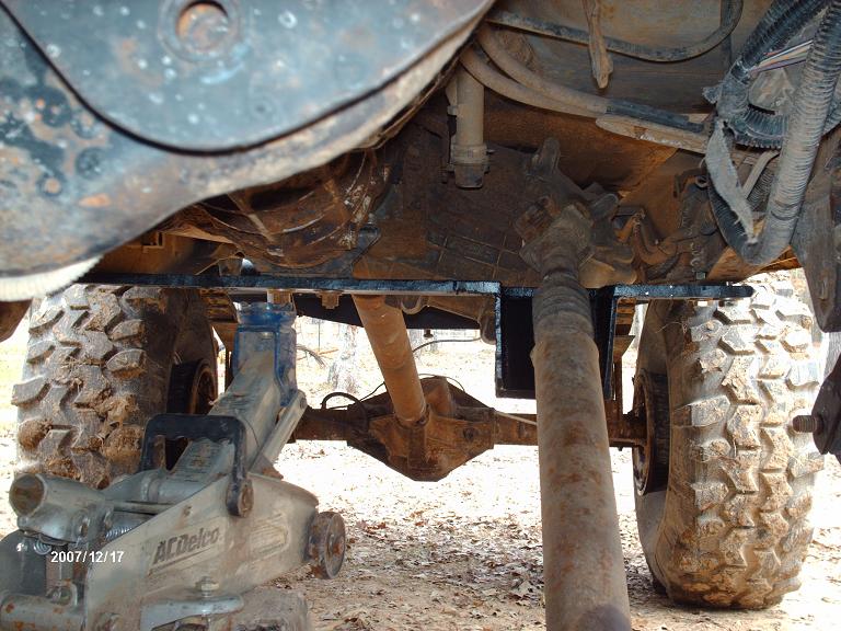 Undercarriage view of a vehicle, showing the transmission, driveshaft, and rear axle, with a floor jack supporting the frame