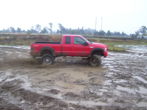 1986 Ford Bronco - A red pickup truck navigates a muddy terrain, with water and dirt splashing around its wheels.