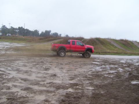 1986 Ford Bronco - A red pickup truck navigates a muddy terrain under a cloudy sky.