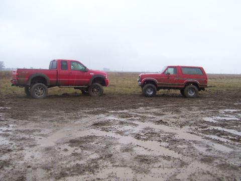 1986 Ford Bronco - Two red trucks, a 1986 Ford Bronco and a pickup, sit in a wet, muddy field under an overcast sky.