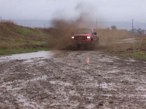 1986 Ford Bronco - A red pickup truck navigates a challenging muddy terrain, creating a large spray of dirt and water.