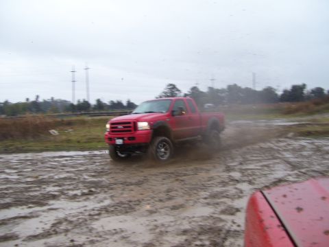 1986 Ford Bronco - Red pickup truck navigating a muddy off-road course under an overcast sky.
