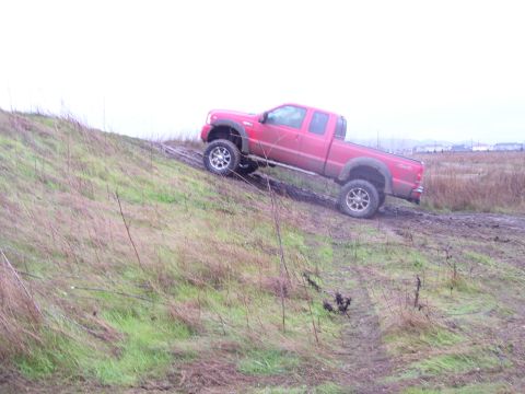 1986 Ford Bronco - A red pickup truck navigates a challenging muddy incline on an overcast day.