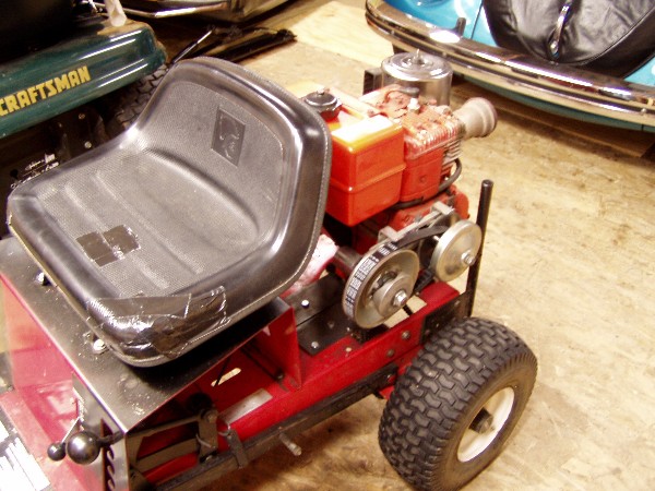 Red lawn tractor frame with black seat and engine components. A green Craftsman mower is to the left