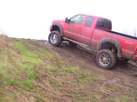 1986 Ford Bronco - A red pickup truck navigates a muddy incline with grass on the side.