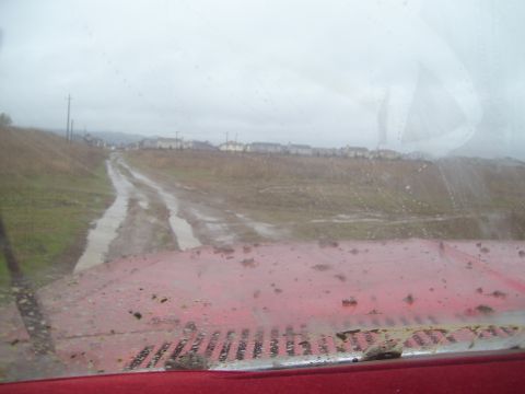 1986 Ford Bronco - View from inside a red vehicle driving on a muddy road with houses in the distance.
