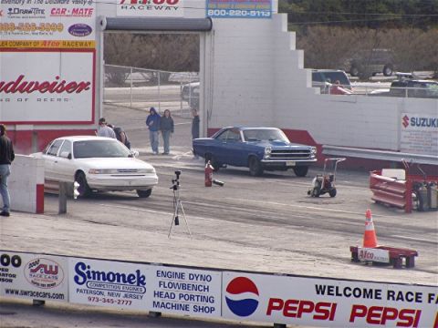 1999 Ford Crown Victoria - A white sedan is positioned at the starting line of a drag strip, with other vehicles and track infrastructure visible in the background.