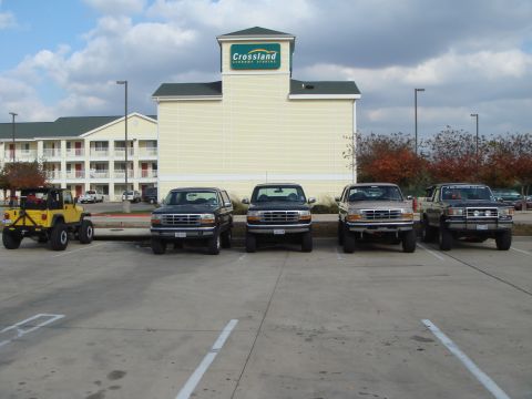 1992 Ford Bronco - A lineup of four 1992 Ford Broncos and a yellow Jeep Wrangler are parked in front of a Crossland Economy Studios hotel.