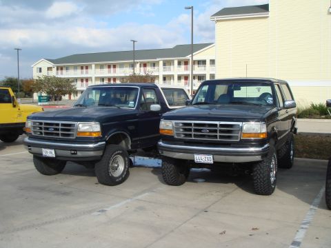 1992 Ford Bronco - Two Ford Broncos are parked side-by-side in a parking lot with a hotel in the background.