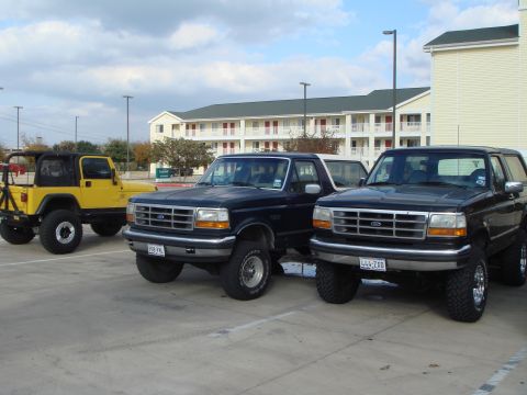 1992 Ford Bronco - Two black Ford Broncos and a yellow Jeep are parked in a lot outside a hotel.