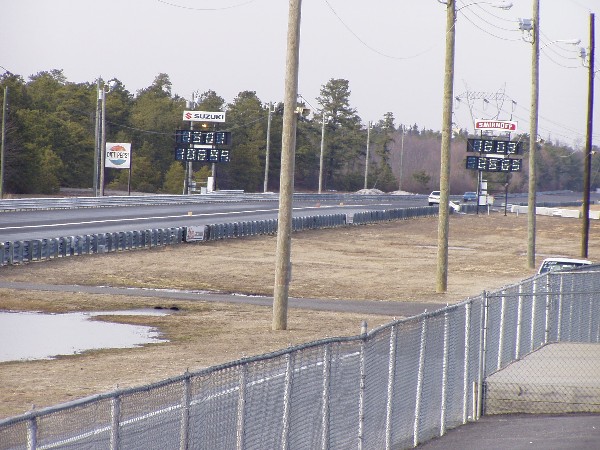 Drag strip with scoreboards and trees in background. A white car is visible on the track