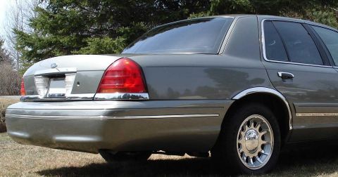 1999 Ford Crown Victoria - Rear three-quarter view of a light olive-gray full-size sedan parked on dry grass in front of evergreens, showing the chrome-trimmed tail l…