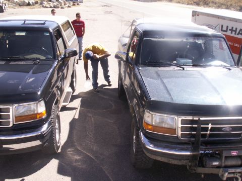 1986 Ford Bronco - Two black Ford F-Series trucks are parked on asphalt, with a person bending over between them.