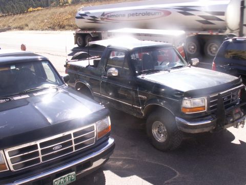 1986 Ford Bronco - A black Ford Bronco is parked next to another vehicle, with a large tanker truck in the background.