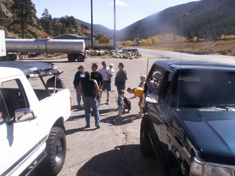 people gathered around a white pickup truck and a dark SUV, with a tanker trailer in the background