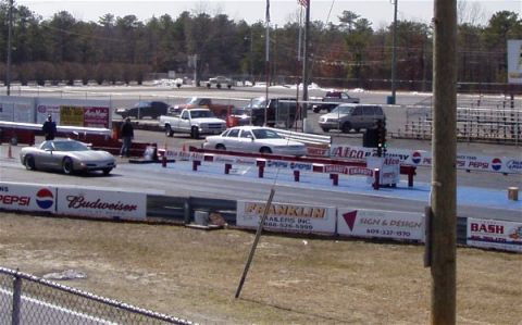 1999 Ford Crown Victoria - A silver sports car is positioned on the drag strip, with other vehicles and spectators visible in the background.