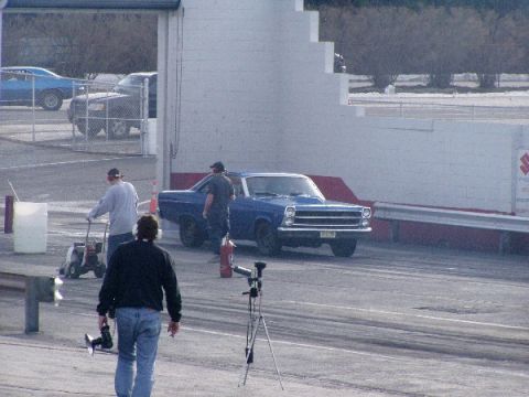 1999 Ford Crown Victoria - A blue pickup truck is seen at a drag strip event with people around.