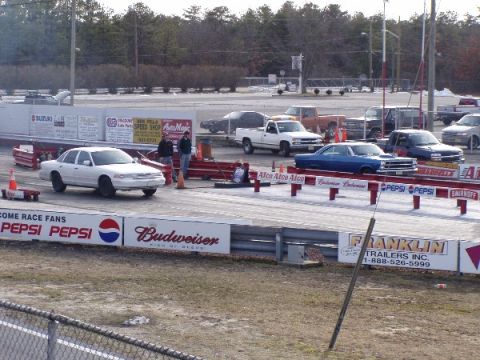 1999 Ford Crown Victoria - A white Ford Crown Victoria is parked on a drag strip with other vehicles and trailers visible in the background.