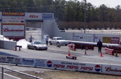 1999 Ford Crown Victoria - A white sedan performs a burnout at a drag strip, with a sports car in the foreground.