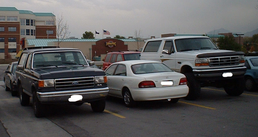White lifted Ford Bronco parked beside a white sedan and dark blue Bronco in a parking lot