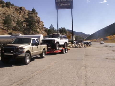 1986 Ford Bronco - A gold pickup truck tows a white Ford Bronco on a trailer past a ski rental sign in a mountainous area.