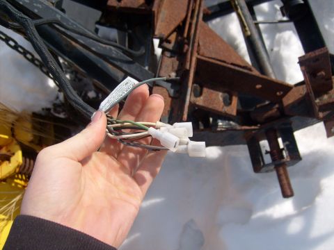 1995 Ford C-350 (Centurion Conversions) - A hand holds a wiring harness connected to a rusty plow mechanism, with snow on the ground.