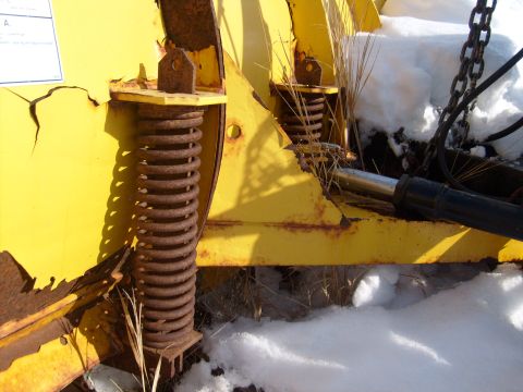1995 Ford C-350 (Centurion Conversions) - Close-up view of the spring mechanism and hydraulic cylinder on a yellow snow plow, partially covered in snow.