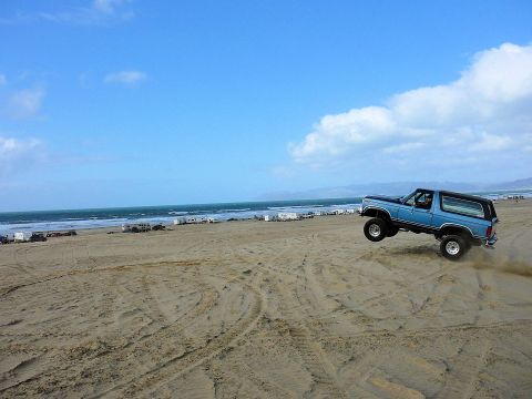 1984 Ford Bronco - A blue SUV is captured mid-air as it drives over sand dunes on a beach with the ocean in the background.