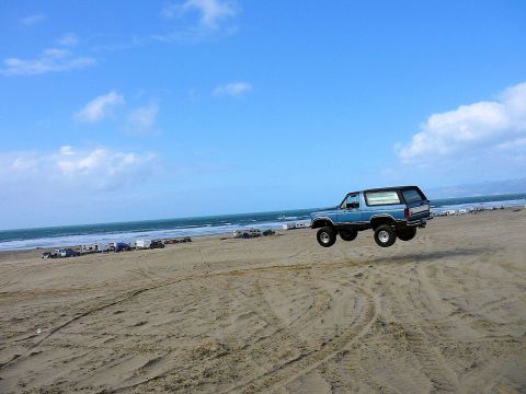 1984 Ford Bronco - A blue SUV is captured mid-air during a jump on a sandy beach with the ocean and other vehicles in the background.