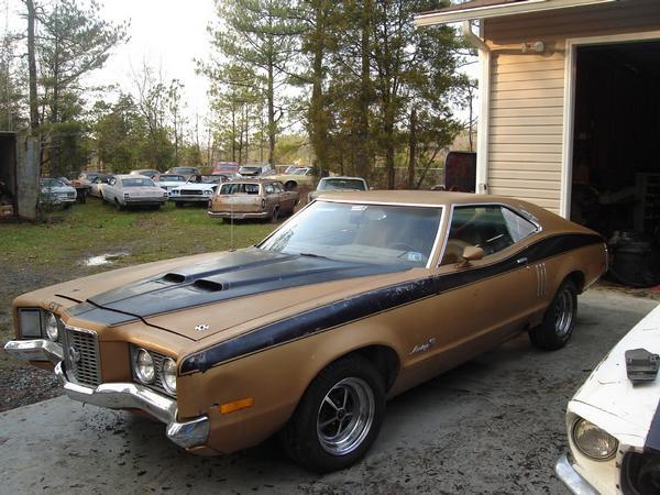 Gold Mercury Montego hardtop with black hood stripes and hood scoops parked by a garage