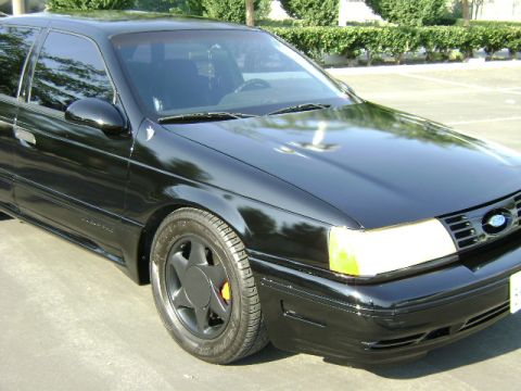 1991 Ford Taurus - A black 1991 Ford Taurus sedan is viewed from the front three-quarter angle, highlighting its distinctive grille and headlights.