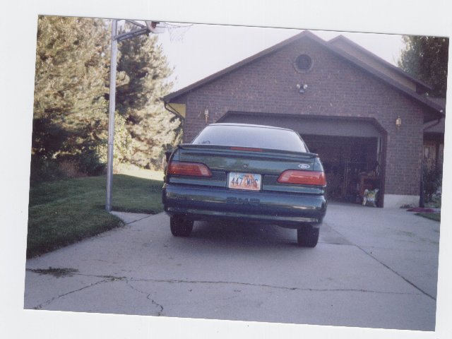 Green Ford sedan rear three-quarter parked in driveway by brick garage, Utah plate