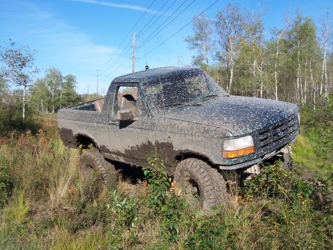 1996 Ford Bronco - A muddy Ford Bronco is stuck off-road, surrounded by vegetation and under power lines.