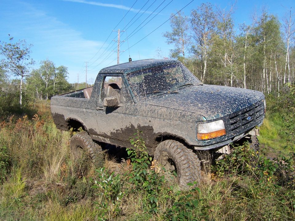 Mud-covered dark green Ford Bronco, 3/4 front view, stuck in tall grass and mud