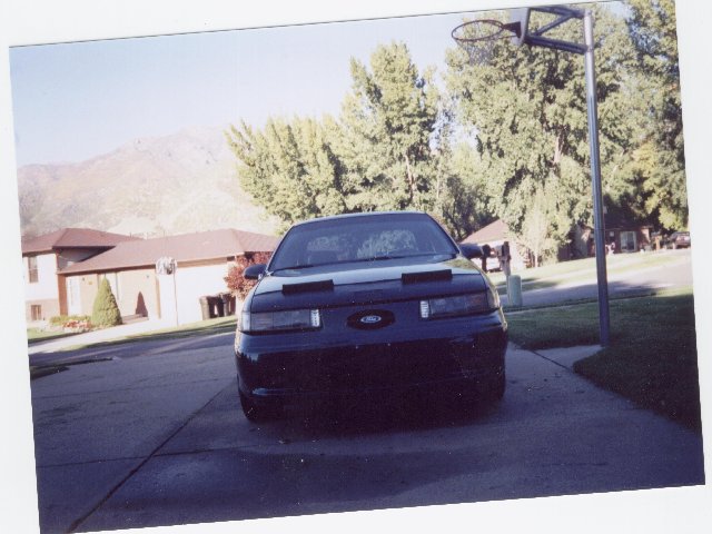 Dark Ford sedan front view in driveway with black bra cover, basketball hoop, mountains