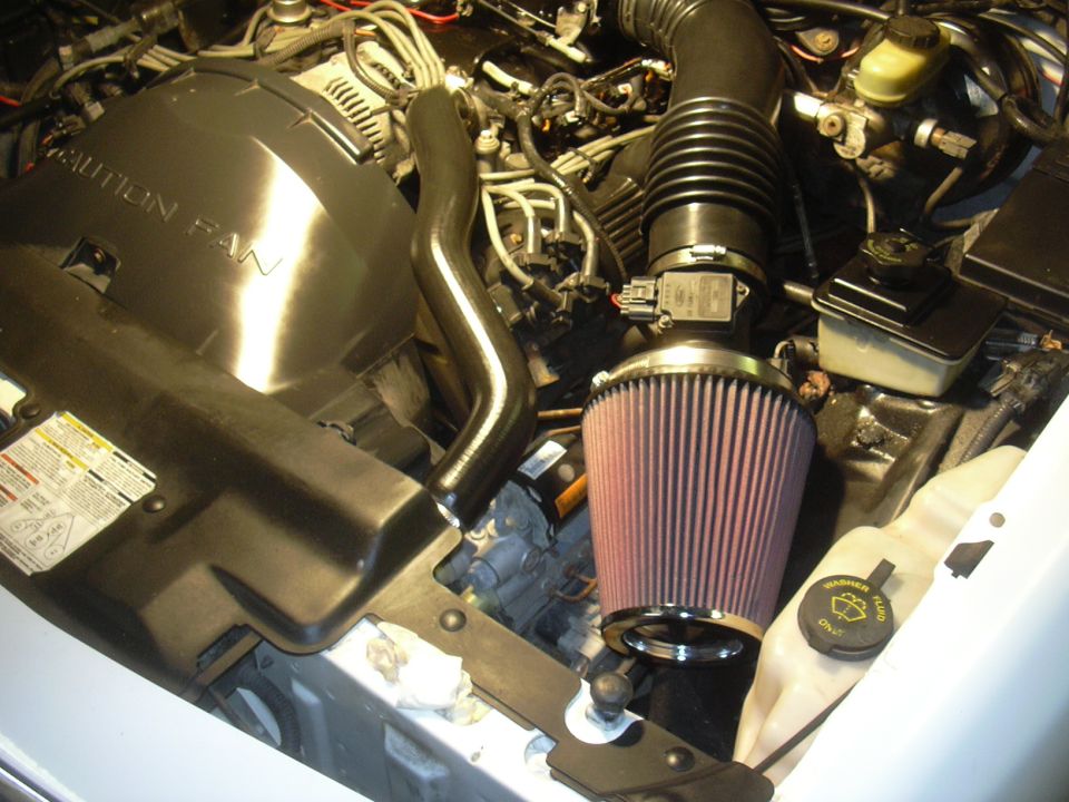 Engine bay with aftermarket air filter and black intake tube. Caution fan cover visible