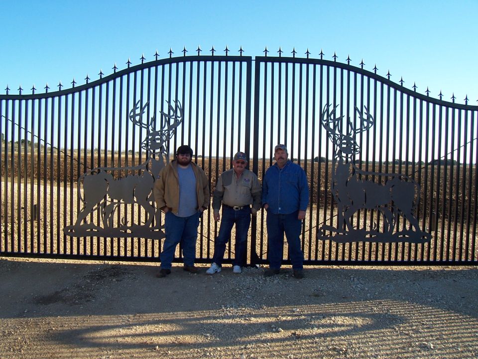 Three men stand before a tall, ornate black metal gate large deer silhouettes on either side