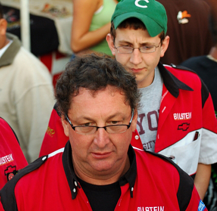 1990 Ford Bronco - Two men in red Bilstein-branded team shirts in a crowd; one older with glasses in front, a younger one in a green cap behind.